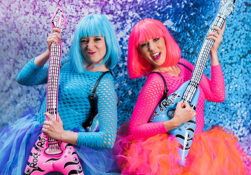 Two women with bright blue and pink wigs, in matching colorful outfits, energetically play inflatable guitars against a vibrant, confetti-filled backdrop.