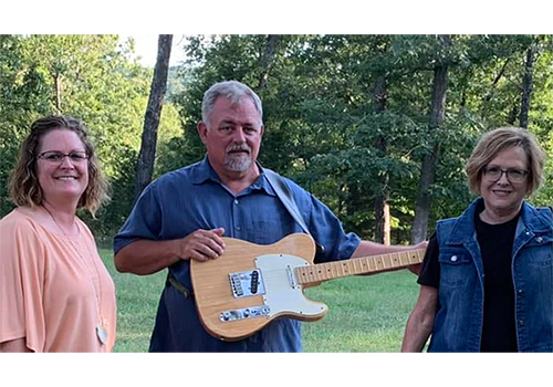 A man with a beige electric guitar stands between two women in a wooded area. They all smile, conveying a relaxed and cheerful mood.