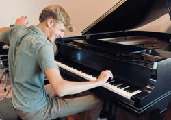 A young man energetically plays a grand piano, smiling with enthusiasm. The room is well-lit, and a drum set is partially visible in the background.