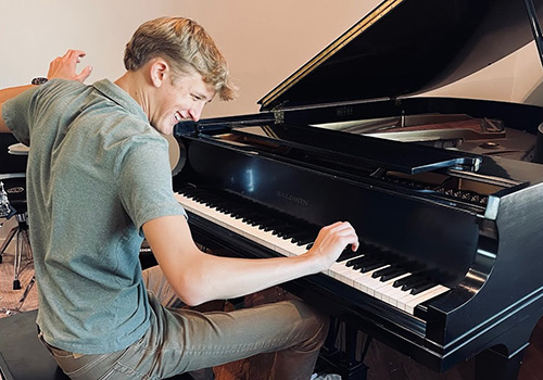 A young man energetically plays a grand piano, smiling with enthusiasm. The room is well-lit, and a drum set is partially visible in the background.