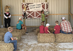 Kids seated on hay bales at a "Story Time with Bart" event. A child stands pointing at a poster, with a costumed character nearby. Casual, engaging atmosphere.