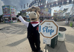A mascot in a vest and tie stands smiling next to a "Selfie Stop" sign at a lively carnival with colorful rides in the background. The atmosphere is festive and cheerful.