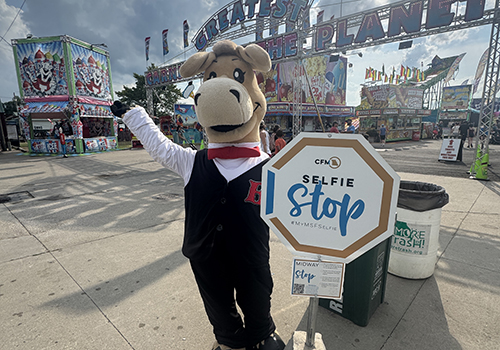 A mascot in a vest and tie stands smiling next to a "Selfie Stop" sign at a lively carnival with colorful rides in the background. The atmosphere is festive and cheerful.