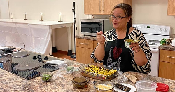 A chef wearing an apron and holding a fork and food by a counter top full of food