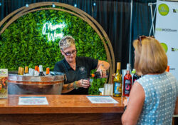 A person with short hair pours wine for a customer at a bar with a green foliage backdrop and "Missouri Wines" neon sign. Several wine bottles are displayed.